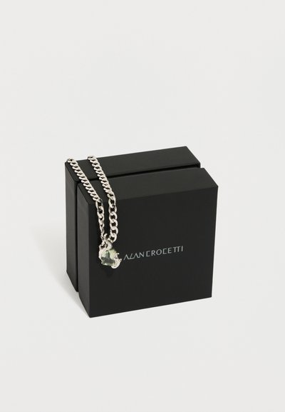 Ķēdes kaklarota ar sudraba saiti un zaļas dārgakmens piekariņu, attēlota kopā ar melnu zīmola kārbu ar minimālu logotipu.