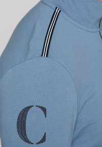 Pullover azzurro chiaro con colletto alto, caratterizzato da un'accenno strisce bianche e nere sulla spalla e una grande lettera "C" nera sulla manica.