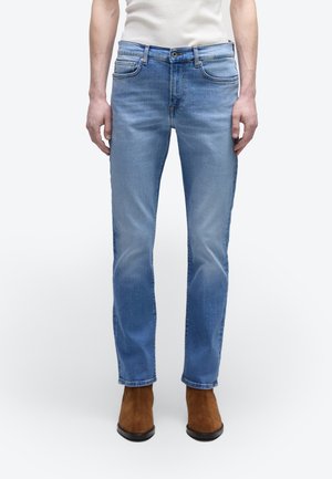 Mann trägt hellblaue Slim-Fit-Jeans, weißes T-Shirt und braune Wildleder-Stiefeletten und steht vor einem einfarbigen Hintergrund.