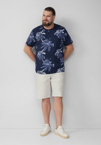 Granatowa koszula z krótkim rękawem w białe wzory palm, zestawiona z jasnymi szortami i białymi butami sportowymi. Strukturalny, swobodny fason.
