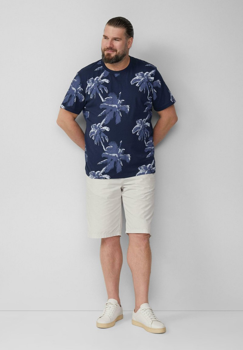 Granatowa koszula z krótkim rękawem w białe wzory palm, zestawiona z jasnymi szortami i białymi butami sportowymi. Strukturalny, swobodny fason.