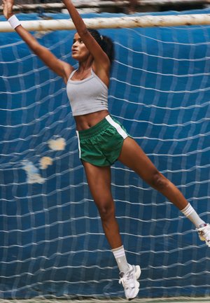 Femme athlétique en short vert et débardeur gris sautant les bras levés devant un filet bleu, semblant attraper ou bloquer quelque chose.