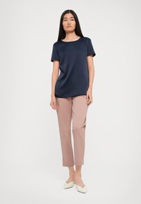 Donna con lunghi capelli scuri indossa una maglietta blu navy a maniche corte, pantaloni rosa chiaro a vita alta, e ballerine bianche su uno sfondo bianco.