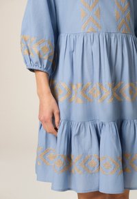 Vestido azul claro con mangas abullonadas, que presenta patrones geométricos bordados en oro cerca del dobladillo y de las muñecas, además de un diseño de volantes en capas.