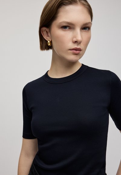 Top tricotat cu nervuri de culoare bleumarin, cu mâneci scurte și guler rotund. Textură netedă cu o siluetă ajustată. Modelul poartă cercei din aur în formă de cerc.