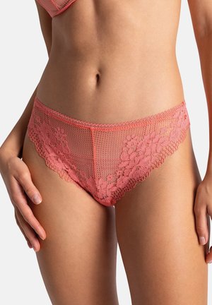 Femme portant une lingerie style bikini en dentelle rose corail et un soutien-gorge assorti, debout, les mains détendues le long du corps, sur fond uni.
