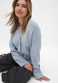 Cardigan lavorato a maglia azzurro chiaro con scollo a V, chiusura con bottoni e polsini a coste. Abbinato a pantaloni scuri e morbidi. Design minimalista.