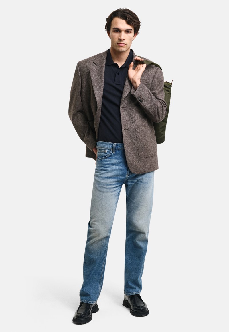 Brauner Wollblazer über einem marineblauen Poloshirt, kombiniert mit hellen Jeans und schwarzen Stiefeln. Das Modell trägt eine grüne Tasche.