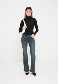 Svart turtleneck, blå flared jeans og svarte spisse ballettsko. Modellen holder en hvit veske og står mot en enkel hvit bakgrunn.