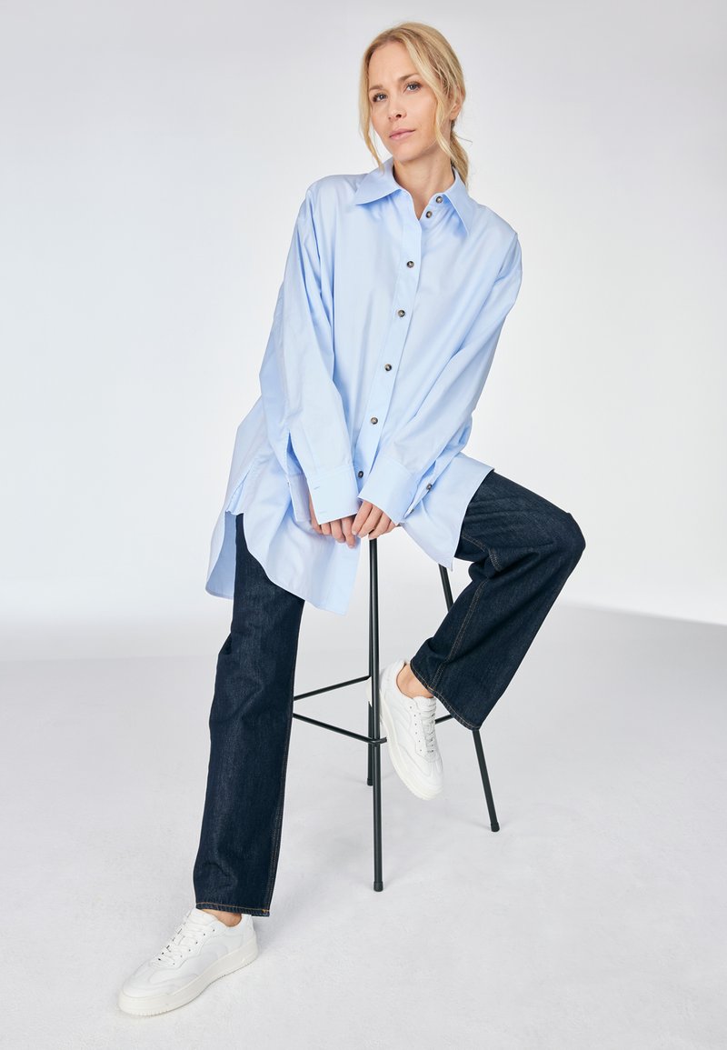 Himmelblaues Oversized-Hemd mit Knöpfen vorne, langen Ärmeln und seitlichen Schlitzen, kombiniert mit dunklen Jeans und weißen Sneakers.