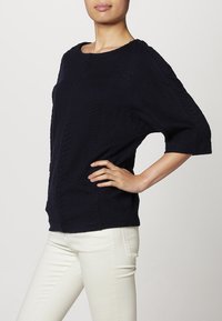Maglione lavorato a maglia blu navy con scollo rotondo, maniche corte e un motivo a zigzag testurizzato. Abbinato a pantaloni attillati di colore chiaro.