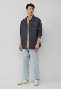 Marineblauwe utilityjack met twee borstzakken, gecombineerd met een beige T-shirt en lichtblauwe relaxed-fit jeans. Schoenen hebben een wit en tan ontwerp.