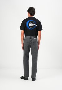 Camiseta negra de manga corta con un gráfico circular azul y texto blanco. Combinada con jeans de mezclilla grises y zapatos negros.
