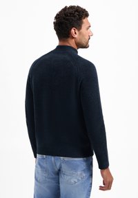 Donkerblauwe gebreide trui met hoge kraag, geribbelde accenten op de schouders en manchetten, en een gestructureerde afwerking. Draagt met lichtblauwe denim jeans.