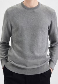 Pull gris en tricot avec col rond, manches longues, texture subtile, et poignets et ourlet côtelés. Porté avec un pantalon foncé.