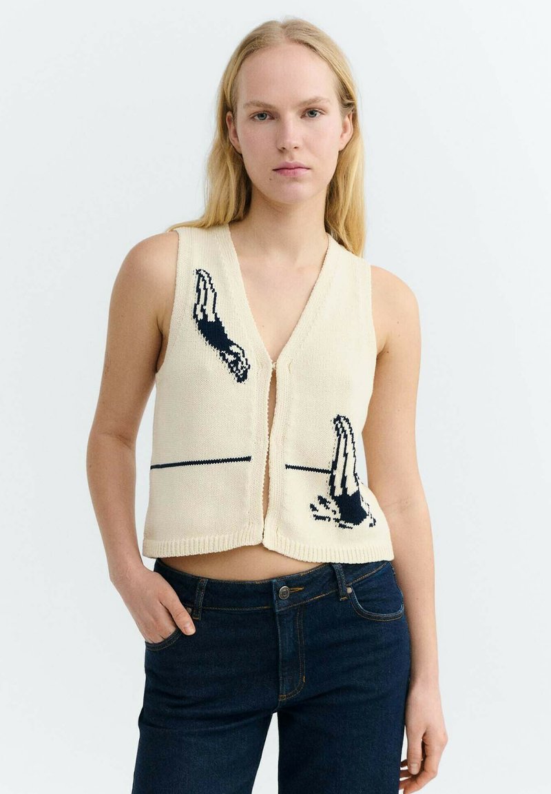 Blonde Frau, die eine ärmellose cremefarbene Weste mit schwarzen Taucher-Mustern und dunkelblaue Jeans trägt, vor einem schlichten weißen Hintergrund stehend.