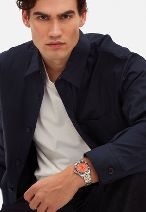 Montre en acier inoxydable avec un cadran orange et un bracelet en maillons argentés, portée sur un poignet. Chemise à boutons bleu marine par-dessus un t-shirt blanc.