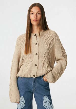 Mujer con un cárdigan de punto grueso beige con botones de madera y vaqueros azules con parches texturizados en azul claro.