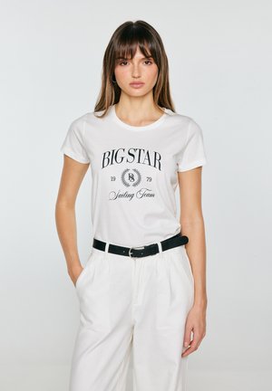 Noor naine sirgete pruunide juustega kannab valget "BIG STAR Sailing Team" t-särki, valgeid pükse ja musta vööd, seisab tavalise tausta ees.