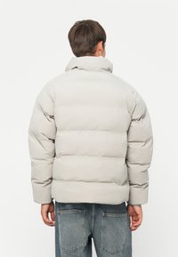 Patterjakke i lysegrå med høj krave, oversized pasform og syede sektioner. Sammen med slidte jeans, der viser baglommer.