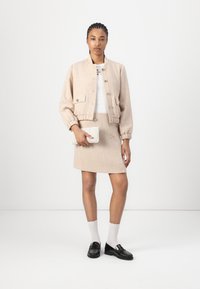 Bež tweed bomber-jakk koos kahe esipocketiga, sobitatud sobiva seeliku juurde. Modell hoiab tekstureeritud valget kotti. Mustad loafers ja valged sokid.
