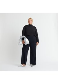Schwarzes Langarmoberteil, hoher Kragen, marineblaue weit geschnittene Hose, silberne metallische Tasche und Slipper. Glatter Stoff, lockere Passform, monochromatisches Outfit.