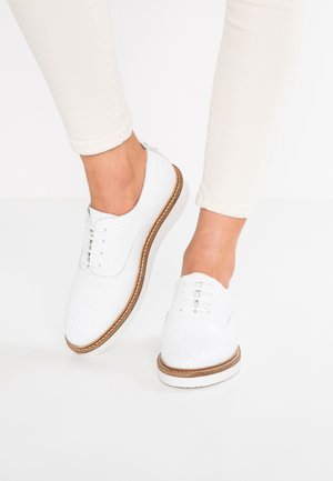 Białe skórzane buty o teksturowanej powierzchni z okrągłym noskiem, pięcioma oczkami i brązową gumową podeszwą, umieszczone na nagich nogach w jasnobeżowych dżinsach.