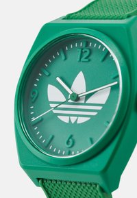 Žalia laikrodžio apyrankė su matinio paviršiaus apdaila, apvaliu korpusu, baltu Adidas logotipu ir žalia tekstūruota juosta. Minimalistinės skaičių ir rodyklių detalės.