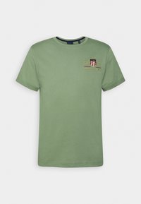 Camiseta de algodón verde con cuello redondo, mangas cortas y un diseño en el pecho que incluye "GANT" y un logo en rojo y dorado.