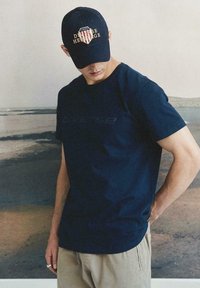 Granatowy bawełniany T-shirt z krótkim rękawem i subtelnym logo. Połączony z khaki szortami i granatową czapką z wyraźnym graficznym napisem.