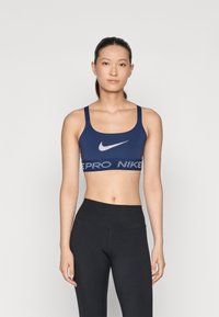 Jūras militārā krāsa sporta krūšturis ar platu joslu, uz kuras ir baltais teksts "PRO NIKE", regulējamām siksnām un centrā esošo balto Nike logo. Gluda, elastīga materiāla.
