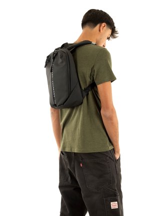 Mochila negra de diseño elegante, con una superficie texturizada y correas ajustables, llevada en la espalda de una persona con una camiseta verde.