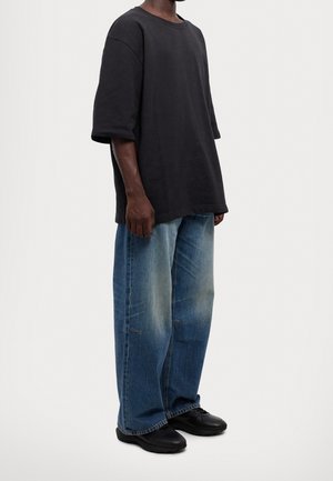 Personne portant un T-shirt noir oversize, un jean bleu ample et des chaussures noires, debout devant un fond clair uni.