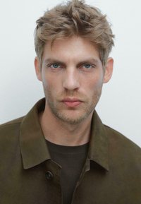 Mann mit hellbraunen Haaren und einem Bart trägt eine dunkelgrüne Wildlederjacke über einem dunklen Hemd, das einen Kragen und auffällige Knöpfe hat.
