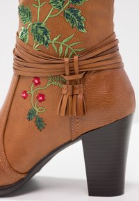 Braune Kunstleder-Ankle Boots mit einem spitzen schwarzen Absatz, verziert mit grüner und roter floralem Stickerei sowie einem geflochtenen Quastenakzent.