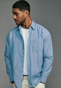 Light blue striped button-up shirt with a chest pocket, worn over a white crewneck t-shirt. Smooth texture and relaxed fit.