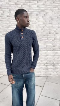 Young man wearing a navy cable knit sweater with wooden buttons and blue jeans, standing with one hand in pocket against a white brick wall.