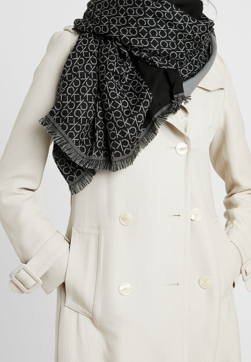 Personne portant un manteau croisé crème avec de gros boutons et une écharpe noire avec des motifs blancs "CK" répétés et des bords frangés.