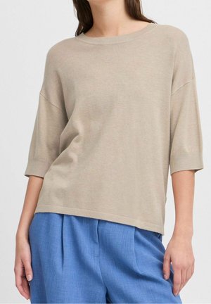 Vrouw draagt een beige loszittend gebreid topje met ellebooglange mouwen en blauwe hoge taille plooibroek.