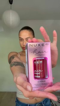 Persoon met getatoeëerde arm houdt een Nuxe haarserumdoos richting de camera vast in een neutrale binnenomgeving.