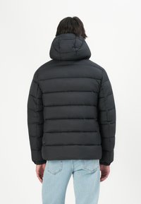 Person iført en sort quilted pufferjakke med hætte, med ryggen til, parret med lyseblå denimjeans mod en ensfarvet hvid baggrund.