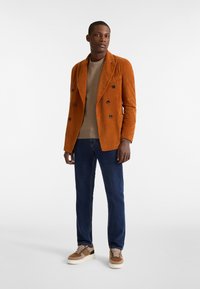 Oranger Cord-Stoff Doppelreiher-Blazer, beiger Pullover, dunkelblaue Jeans und braune Sneakers mit gemusterten Akzenten. Glatte Texturen wahrgenommen.