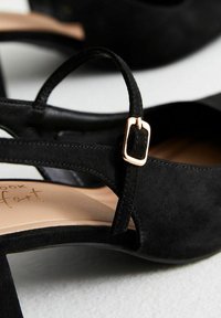 Sandalias de tacón alto de ante negro con una correa curvada y detalle de hebilla de metal dorado. Presenta una plantilla beige suave y un diseño de punta abierta.