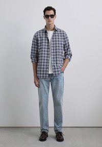 Camisa de cuadro en azul marino y blanco, camiseta blanca, jeans de un azul claro y zapatos oscuros de cordones; el modelo está de pie con las manos en los bolsillos.