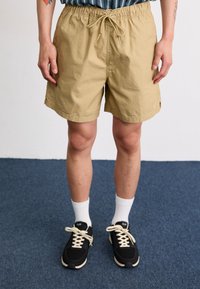 Bermudas bejes com um cós elástico, fecho de cordão e bolsos laterais. Em combinação com ténis pretos e meias brancas de cano curto.