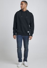Sudadera negra con bolsillo frontal tipo canguro, capucha con cordón y puños de canalé, combinada con jeans azules y zapatillas blancas. Diseño simple.