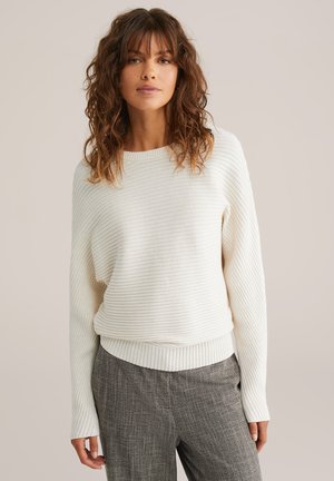Pulover alb cu un model striat, cu mâneci lungi și guler rotund, având o croială lejeră și o țesătură texturată. Combinate cu pantaloni gri cu model.