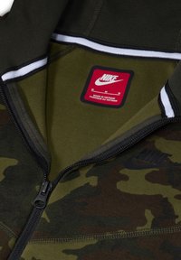 Felpa con zip in camo verde e marrone con accenti neri, caratterizzata da un bordo a righe bianche e un'etichetta rossa Nike sul colletto interno.
