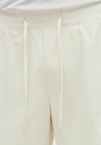 Primer plano de pantalones de tela texturizada color crema con una pretina elástica y dos cordones colgando al frente.