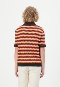 Korte erm polo-genser med strikk, med et chevronmønster i oransje, rødt og hvitt, samt svart krage og mansjetter. Glatt tekstur.